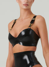 A black latex bra with gold suspender clasps and rings, featuring adjustable shoulder straps and fastened at the back. The model is wearing the bra with a matching latex lingerie set.