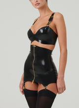 A model wears a black latex rubber bra and matching girdle with a gold metal front zip. Bra and Girdle feature gold hardware including gold suspender clips. Also wearing black opaque stockings.