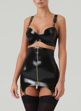 A model wears a black latex rubber bra and matching girdle with a gold metal front zip. Bra and Girdle feature gold hardware including gold suspender clips. Also wearing black opaque stockings.