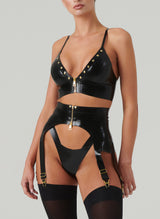 A model wearing a black latex lingerie set and black stockings. The set includes gold studded bra and suspender belt, both with gold front zips, and matching latex thong.