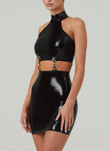A model wearing a shiny black latex mini dress with halter neck collar, and cutout at the waist that fastens with gold hardware.