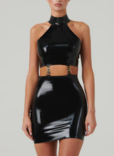 A model wearing a shiny black latex mini dress with halter neck collar, and cutout at the waist that fastens with gold hardware.