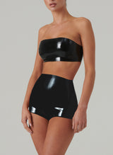 A model on a plain background wears shiny black latex boob tube and high waisted hot pants.