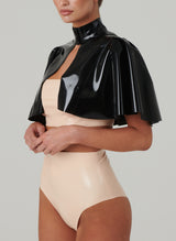 A person wearing a black latex mini cape with flared inserts, fastened with press studs, over a pale peach latex boob tube and big knickers.