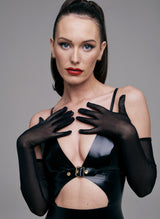 Close up of a model wearing a black latex dress with a front cutout and gold buckle detail, with adjustable shoulder straps. Black mesh long gloves are also worn.