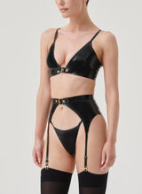 Side view of a model wearing a black latex lingerie set with gold buckles, including a bra, suspender belt and G-string, all by William Wilde. Model also wears black nylon stockings.