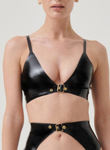 A model wearing a black latex bra with a gold buckle at the front, with skinny adjustable shoulder straps. Matching latex suspender belt also in shot, all by William Wilde.