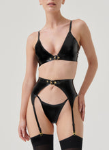 A model wearing a black latex lingerie set with gold buckles, including a bra, suspender belt and G-string, all by William Wilde. Model also wears black nylon stockings.