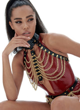 A model wearing a plum latex bodysuit, underneath black leather chest harness and belt, with gold rings and draped gold chain.