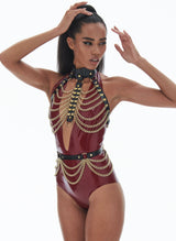 A model wearing a plum latex bodysuit, underneath black leather chest harness and belt, with gold rings and draped gold chain.