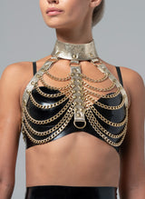 A gold leather harness with scalloped chain detailing at the bust, featuring adjustable straps and chains. Worn over a black latex bra.