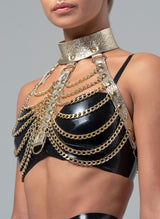 A gold leather harness with scalloped chain detailing at the bust, featuring adjustable straps and chains. Worn over a black latex bra.