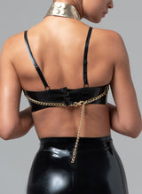 Back view of a gold leather harness with scalloped chain detailing at the bust, featuring adjustable straps and chains. Worn over a black latex bra.