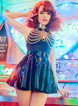 A model with ginger hair wears a gold leather and chain harness over a black mesh top and black latex skating skirt, on a neon background.