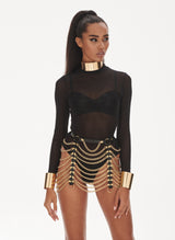 A model wears a black mesh bodysuit with long sleeves under black leather harness accessories with gold studs and chains attached.