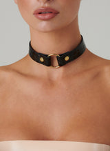 A close-up image of a person wearing a black leather choker with a gold metal O-ring and rivet details.