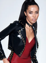 A model wearing a black latex biker jacket with gold studs, over a plum latex dress and black leather choker with gold O-ring at centre.