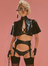 A model wearing a black latex lingerie set, short cape and lace eye mask, on a pink background. The set includes a bra with crossover straps at front, suspender belt and matching latex hipster briefs. Worn here with black opaque stockings. the model carries a riding crop. 