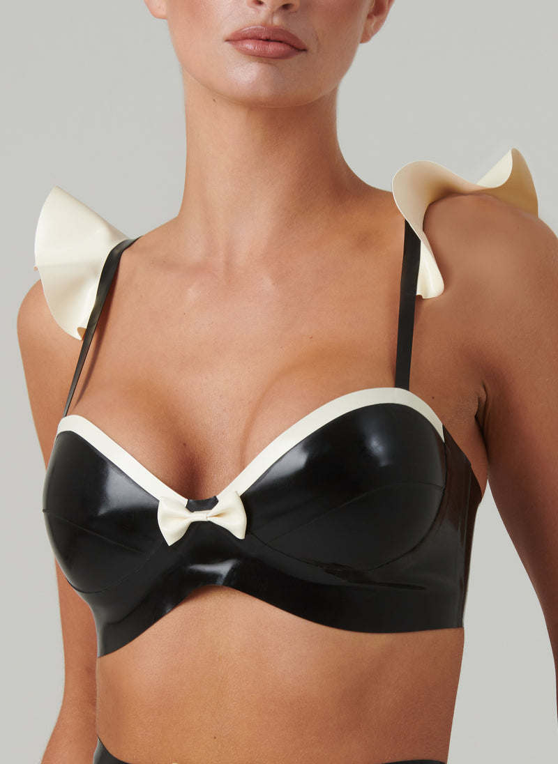 A black latex bra with shoulder frill, mini bow at front and trim in white latex, worn by a model on a plain background.