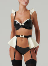 Black and white latex rubber lingerie set including a bra and suspender belt with frills and bows, and matching hipster briefs. Worn by a model also wearing black stockings, on a light grey background.