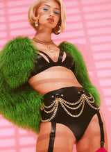 Woman wearing a green fur cropped jacket, black latex bra, and high-waisted black latex knickers with gold chain belt, against a pink background.