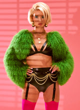 Woman wearing a green fur cropped jacket, black latex bra, and high-waisted black latex knickers with gold chain belt, against a pink background.